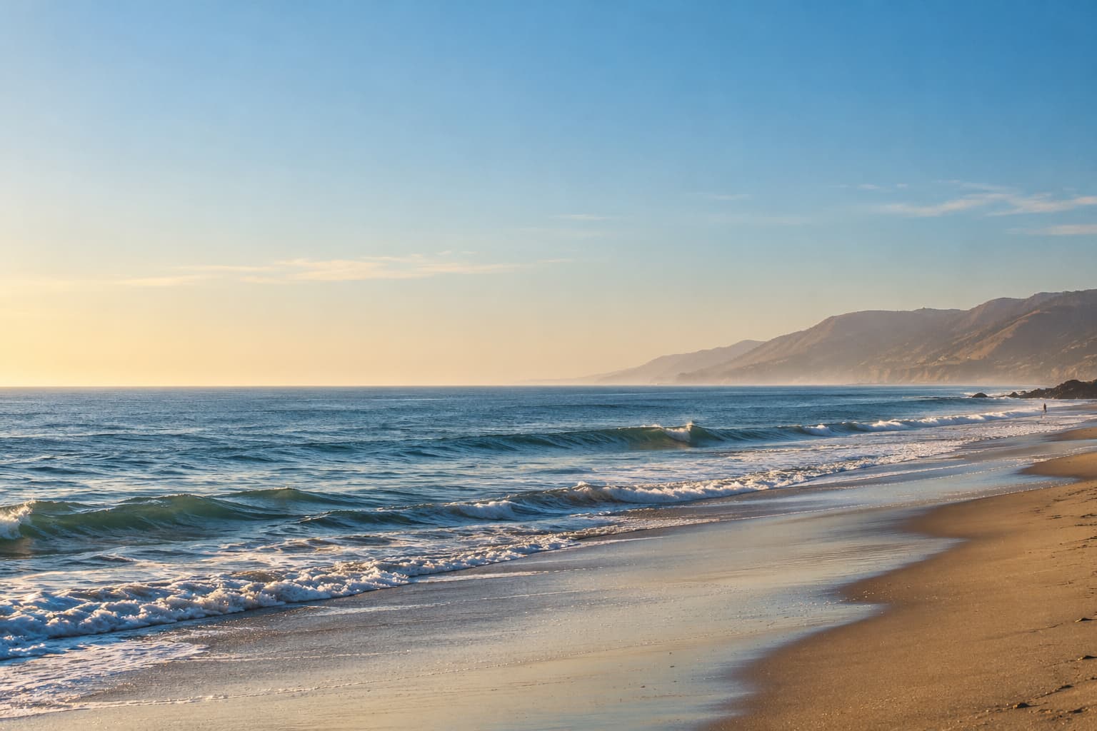 Zuma Beach in Malibu on a clear morning, with waves rolling onto a wide stretch of sand and coastal hills fading into the distance.